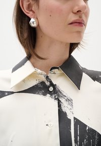 White blouse with bold black abstract patterns, button-up front, and a classic collar. Model wears a shiny silver earring.