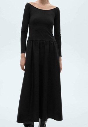 Kobieta ubrana w długą czarną suknię z długimi rękawami i odsłoniętymi ramionami, stojąca na tle jednolitego jasnego tła.