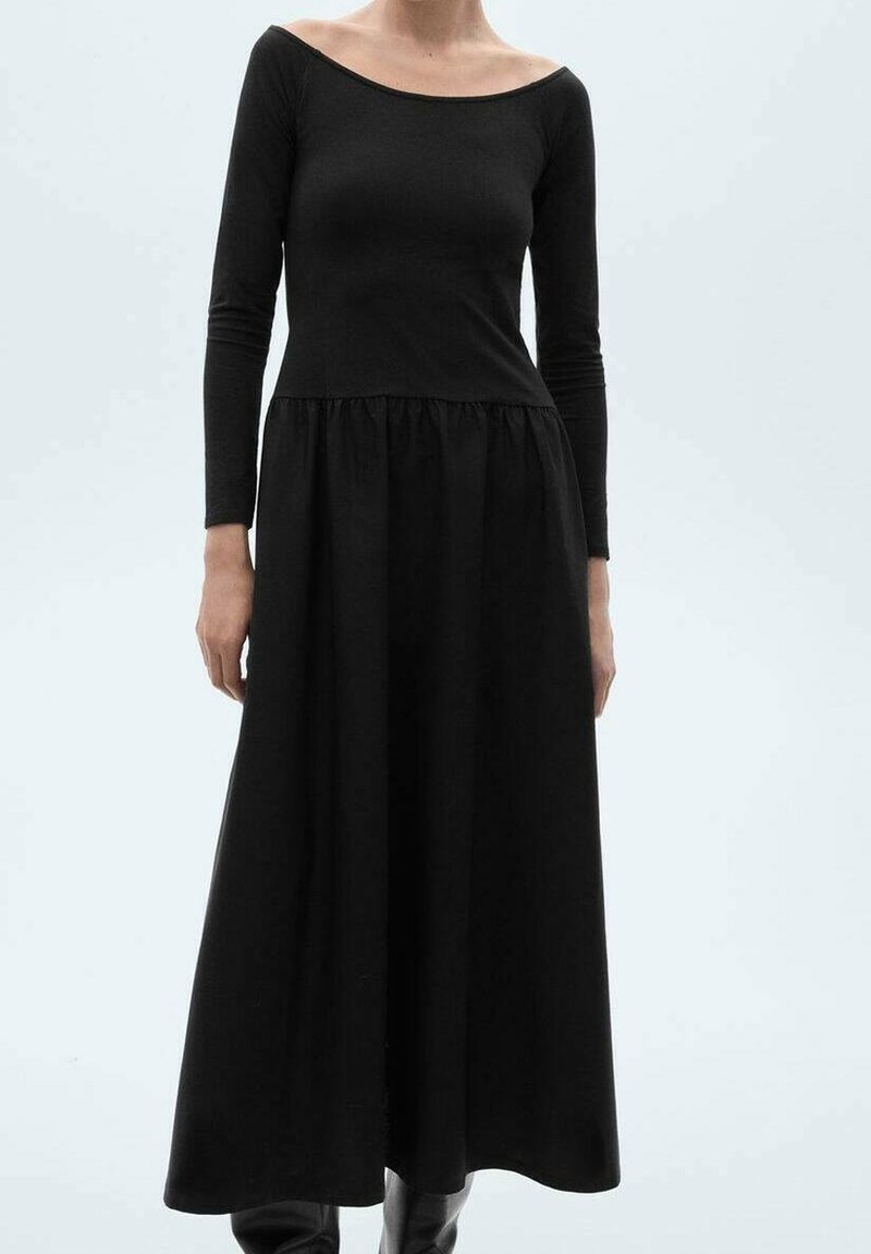 Femme portant une longue robe noire à manches longues et col dégagé, debout devant un fond clair uni.