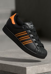 Svart lær sneakers med oransje striper, snøring og gummisåle. Har en strukturert tåhette og logo på tungen.