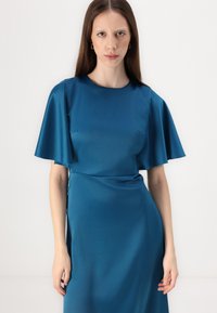 Robe en satin couleur sarcelle avec des manches courtes à volants, un col rond et une silhouette ajustée. Texture lisse avec un léger éclat.