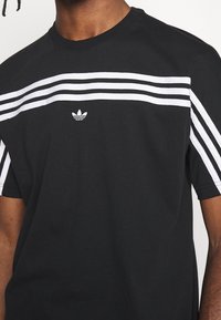 Camiseta de algodón negra con rayas horizontales blancas en el pecho y los hombros, con un pequeño logo bordado en el centro.