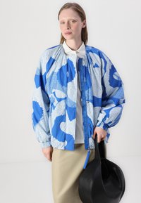 Jaqueta bomber azul com padrão e mangas bufantes, fechada na frente, combinada com uma saia bege clara e uma bolsa de couro preta.