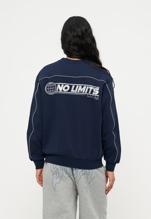 Person med langt, mørkt hår iført en marineblå sweatshirt med teksten "NO LIMITS" og globuslogo på ryggen samt grå joggingbukser med blå grafisk tekst.