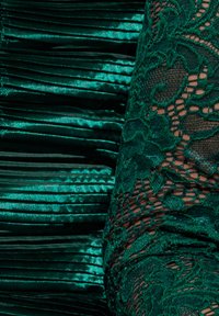 Vihreä mekko, jossa satiinipleettejä ja monimutkaista pitsikuviointia. Pleettikangas on kiiltäväpintainen, ja pitsi on läpinäkyvää kukkakuvioineen.