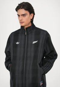 Casaco escocês preto com colarinho alto, apresentando logótipos bordados em branco no peito. Tecido texturizado com fecho de correr e bolsos laterais.