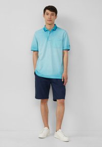 Jasnoniebieska koszulka polo z kieszonką i niebieskimi wykończeniami, w zestawie z granatowymi szortami i białymi trampkami, na jednolitym tle.