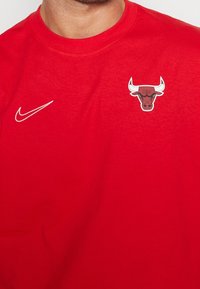 Hombre vestido con una camisa roja que tiene el logo blanco de Nike en el lado izquierdo del pecho y el logo de la cabeza de toro de los Chicago Bulls en el lado derecho del pecho.