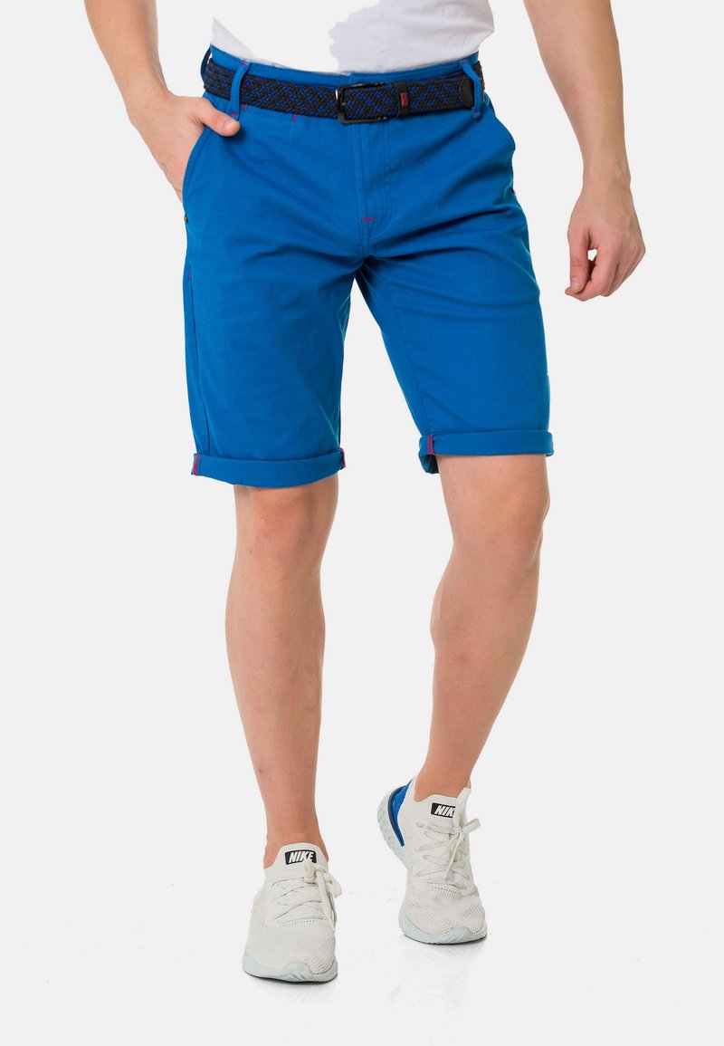 Blaue Baumwollshorts mit umgeschlagenem Saum, kontrastierenden roten Nähten und einem gewebten schwarzen Gürtel. Kombiniert mit weißen Sportschuhen.