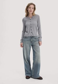 Grijze langemouwen cardigan met knopen aan de voorkant, gecombineerd met loszittende, lichtblauwe jeans met een vervaagde wassing en wijde pijpen. Donkere schoenen maken de outfit compleet.