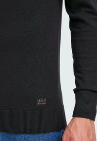 Svart stickad tröja med ribbade muddar och nederkant. Har en brun etikett med texten "ISOLID Denmark Since 1999" nära nederkanten.