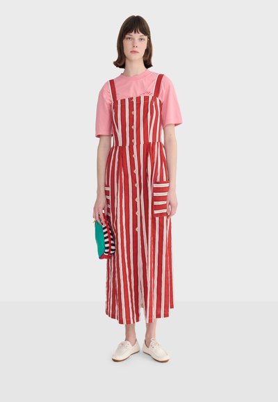 Jeune femme portant une robe rayée verticale rouge et blanche par-dessus un t-shirt rose, tenant une petite pochette multicolore, debout sur un fond uni.