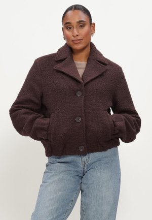Woman with hair pulled back wears dark brown buttoned wool jacket and light blue jeans, standing with hands in pockets against white background.