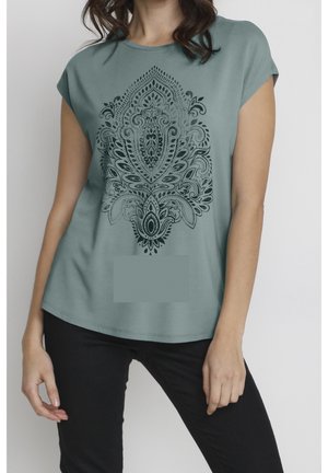 Femme portant une chemise à manches courtes vert mousse de mer avec un motif floral paisley noir complexe, associée à un pantalon noir.