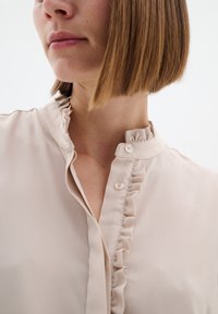 Blusa beige con colletto arricciato e chiusura con bottoni, caratterizzata da un tessuto liscio e texturizzato. Inquadratura ravvicinata sulla zona del collo e parte superiore del corpo.