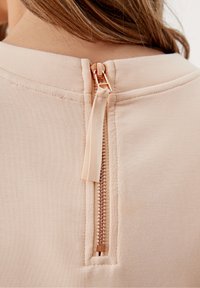 Merch látkový oděv v broskvové barvě s krátkým zipem na zádech. Zip má povrch z růžového zlata a odpovídající textilní táhlo.