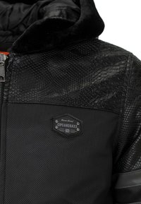 Black jacket featuring textured reptile-like material on one shoulder, smooth fabric on the other, and a logo badge with silver accents.