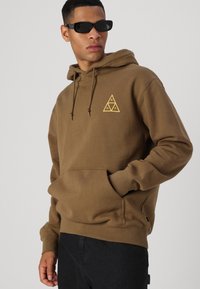 Brauner Hoodie mit einem dreieckigen gelben Logo auf der Brust, Kapuze mit Kordelzug, Kängurutasche und gerippten Bündchen, kombiniert mit schwarzen Sonnenbrillen.