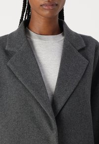 Cappotto in misto lana grigio con rever a punta, apertura frontale e texture morbida, indossato sopra un maglione grigio chiaro con scollatura a costine.