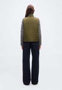 Γυναίκα που φοράει ελιά πράσινο καπιτονέ γιλέκο, γκρι πουλόβερ και σκούρο τζιν με φαρδιές παντελόνες, στέκεται με την πλάτη προς την κάμερα σε απλό φόντο.