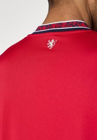 Κόκκινο πλεκτό πόλο με σχέδιο στο γιακά και λευκό έμβλημα λιονταριού στην πλάτη. Το ύφασμα έχει υφή και εφαρμοστή γραμμή.