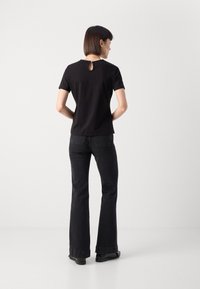 Sort kortærmet top med en detaljeret nøglehul i ryggen, parret med sorte flared jeans og sorte sko, sat mod en ensfarvet hvid baggrund.