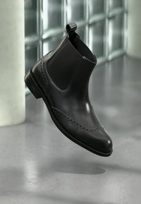 Μαύρο δερμάτινο μποτάκι Chelsea με διακοσμήσεις brogue, ελαστικές πλαϊνές πάνελ και θηλιά για να το τραβάς. Ομαλή υφή και κομψός σχεδιασμός αναδεικνύουν το σχήμα του.