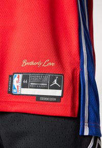 Maglia da basket rossa con texture in rete. Presenta strisce laterali blu e bianche e pantaloni neri. Include la scritta "Brotherly Love" e etichette.
