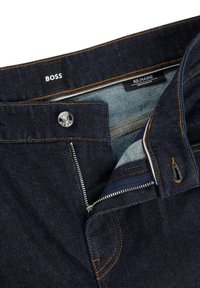 Dark blue denim jeans featuring orange stitching, a front zipper, a snap closure, and brand labels inside the waistband. Regular fit design.