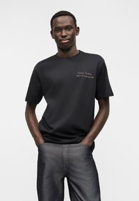 Μαύρο βαμβακερό μπλουζάκι με κοντά μανίκια, με χρυσή επιγραφή στο στήθος. Συνδυασμένο με σκούρο τζιν παντελόνι, το μοντέλο στέκεται με τα χέρια στις τσέπες.