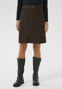Jupe en velours côtelé marron avec deux poches avant, de longueur mi-mollet. Associée à des bottes en cuir noires jusqu'aux genoux avec boucles.