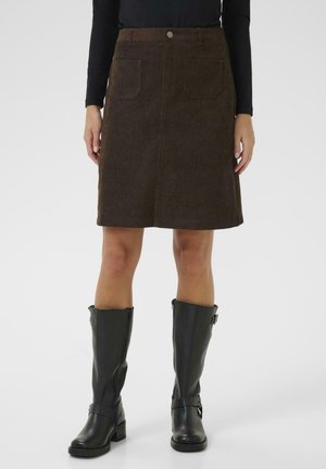 Jupe en velours côtelé marron avec deux poches avant, de longueur mi-mollet. Associée à des bottes en cuir noires jusqu'aux genoux avec boucles.