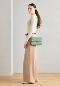 Woman with long curly hair wearing beige ribbed top, wide-leg tan pants, black belt, cream shoes, and green shoulder bag against plain background.