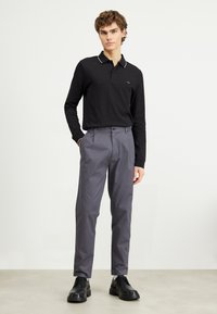 Polo a maniche lunghe nera con colletto bianco, abbinato a pantaloni grigi. Scarpe nere con suola spessa. Design minimalista e stile aderente.