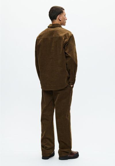Homme debout, de dos, portant une veste en daim marron, un pantalon assorti et des chaussures marron foncé sur un fond uni clair.