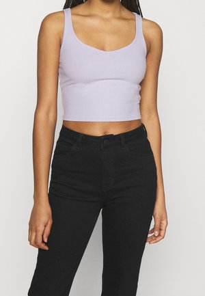 Frau mit ärmellosem fliederfarbenem Crop-Top und hochtaillierter schwarzer Hose, steht mit entspannten Armen vor neutralem Hintergrund.