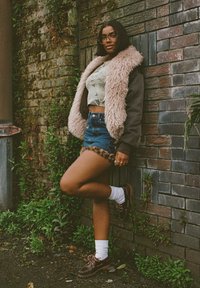 Fuzzy beige collar jacket over a graphic tee, paired with denim shorts featuring leopard trim, white socks, and brown shoes. Grey brick background.