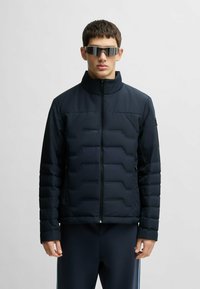 Marinblå puffjacka med hög krage, fyrkantig quiltad mönster och dragkedja fram. Slät tyg med matt finish; bärs med matchande mörka träningsbyxor.