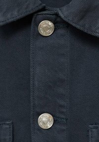 Tmavomodrá džínová košeľa s golierom a dvomi striebornými patentkami, vyrobená z textúrovanej látky s viditeľnými švami.