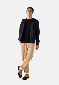 Giacca zip-up blu navy sopra una maglietta nera, pantaloni beige e scarpe marroni. La giacca presenta un cappuccio e polsini elasticizzati; texture del tessuto liscia.