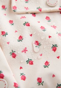 Nærbillede af cremefarvet stof med pink rose og hjerteprint, dækket af vanddråber, der viser dets vandafvisende tekstur.