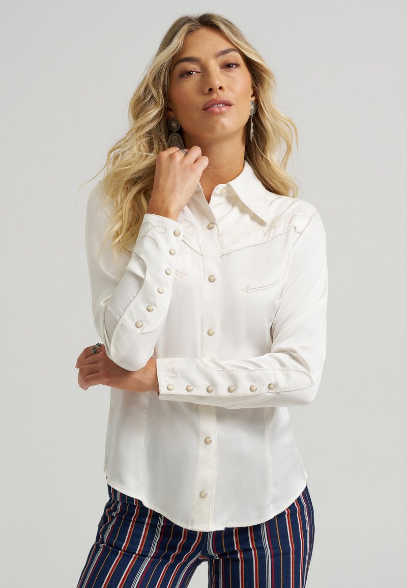 White long-sleeve shirt with a collar, decorative stitching, and pearl-button accents. Styled with vertically striped pants for contrast.