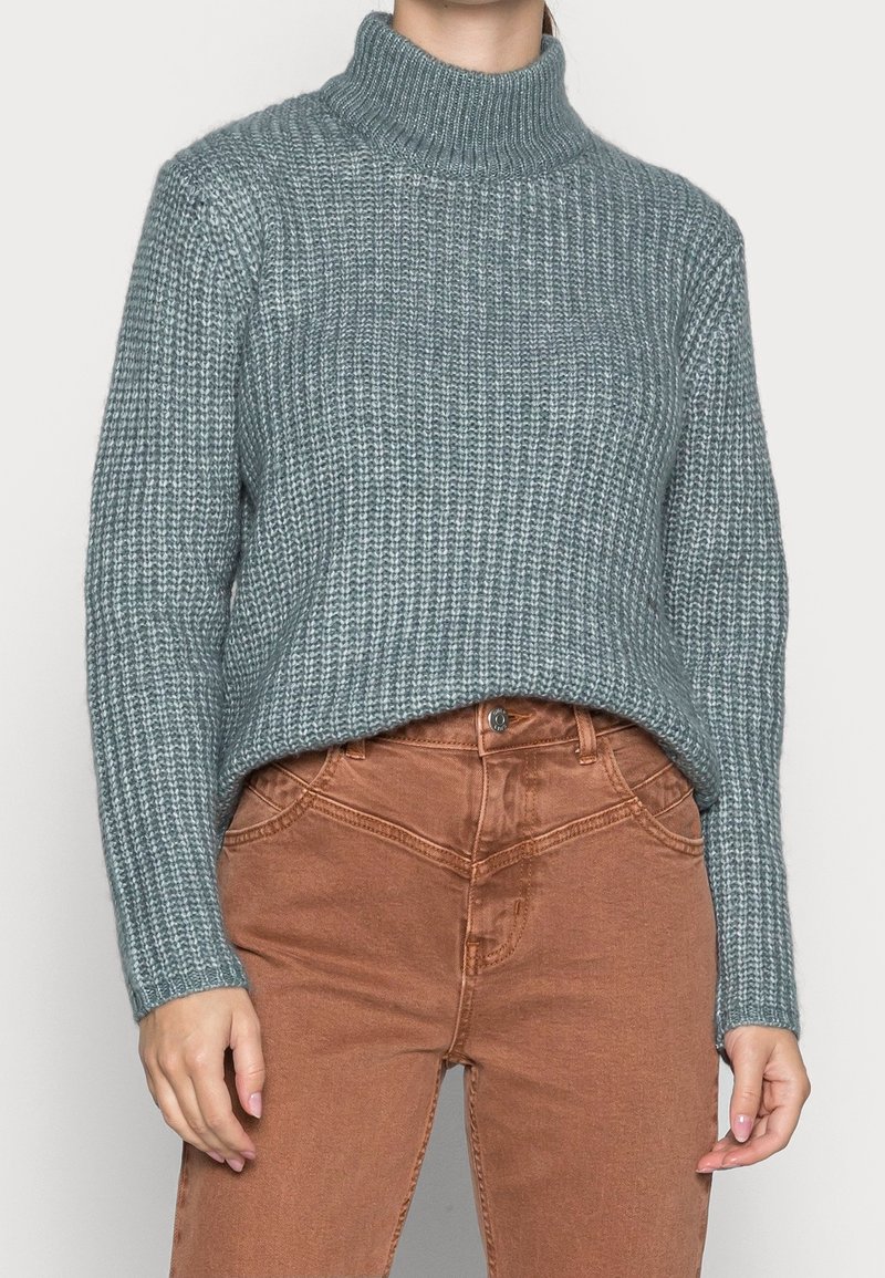 Pull en tricot côtelé turquoise avec un col montant, associé à un jean marron taille haute. Le pull présente une coupe décontractée et un tissu texturé.