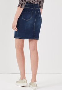 Jupe en denim bleu foncé avec des coutures claires, design ajusté, longueur mi-cuisse, dotée de deux poches arrière et associée à des chaussures claires.