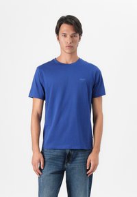 T-shirt blu a girocollo in cotone, con maniche corte e un piccolo logo sul lato sinistro del petto. Indossato con jeans blu.