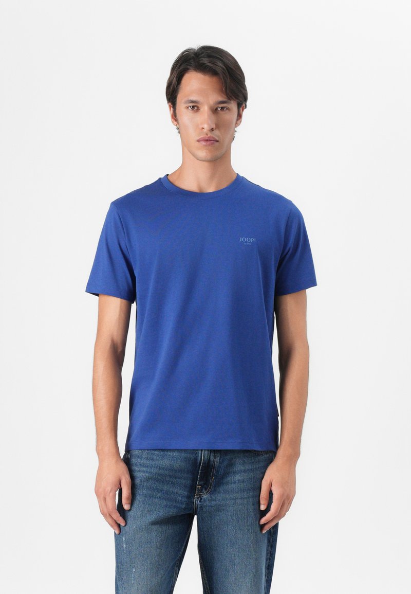 T-shirt blu a girocollo in cotone, con maniche corte e un piccolo logo sul lato sinistro del petto. Indossato con jeans blu.