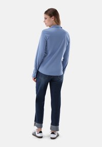 Blaues Langarmshirt mit Kragen, kombiniert mit dunkelblauben Jeans mit hochgekrempelten Bündchen. Das Modell trägt schwarze und weiße Sneaker.