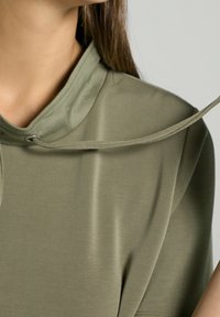 Top verde oliva realizzato in tessuto liscio, con un colletto alto e un sottile nastro regolabile all'uscita. Maniche corte con rifinitura pulita.