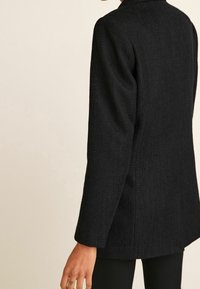 Personne portant un blazer noir texturé à manches longues avec une coupe droite, se tenant devant un fond beige neutre.