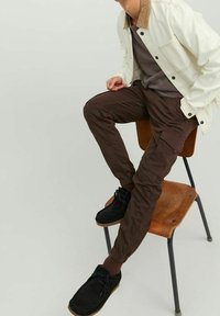 Pantaloni cargo marroni con polsini elasticizzati, abbinati a una maglia grigia e a una giacca di jeans bianca. Scarpe nere su una sedia di legno, su uno sfondo chiaro.